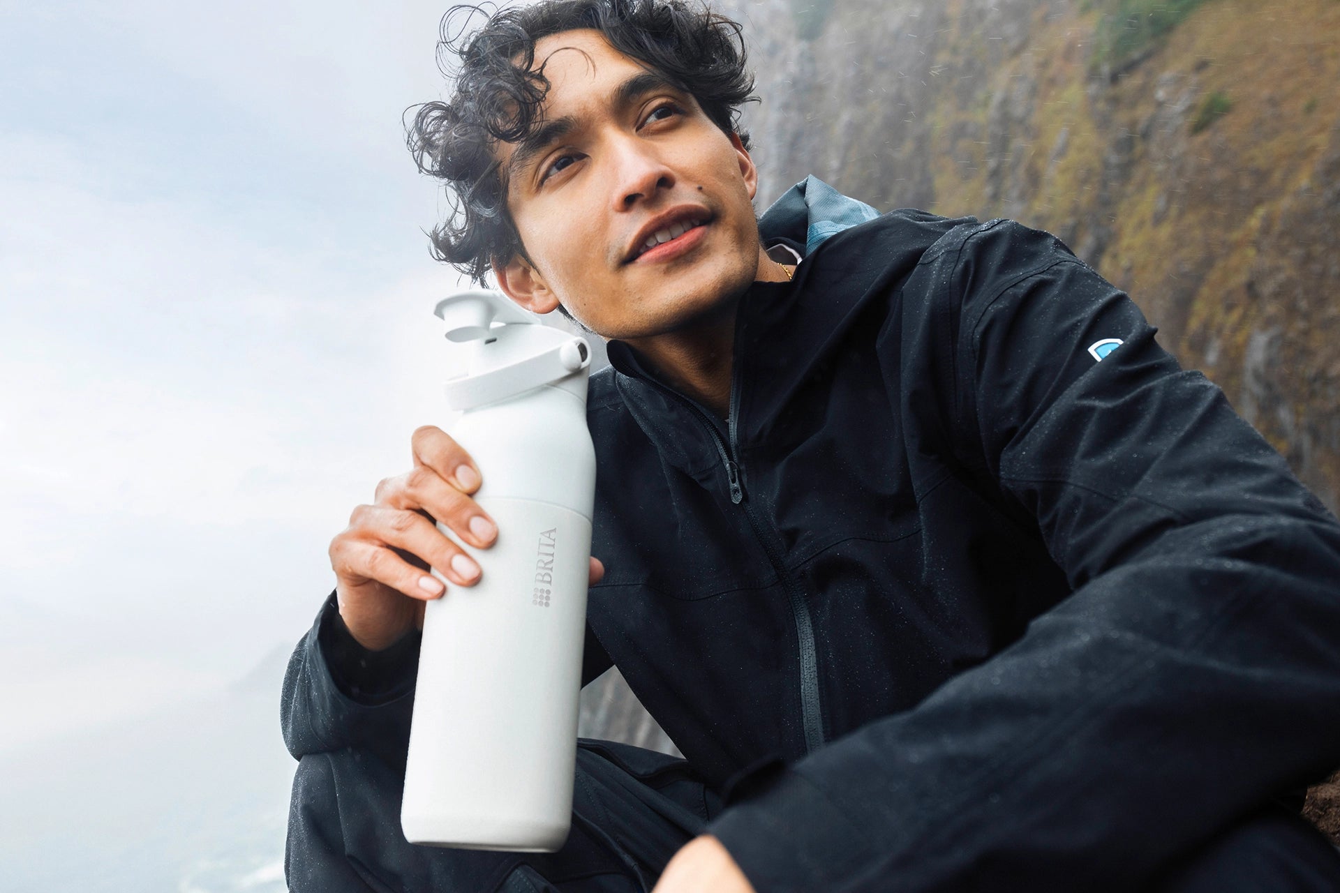 White BRITA LARQ iQ bottle held by a person sitting outdoors in a natural setting.