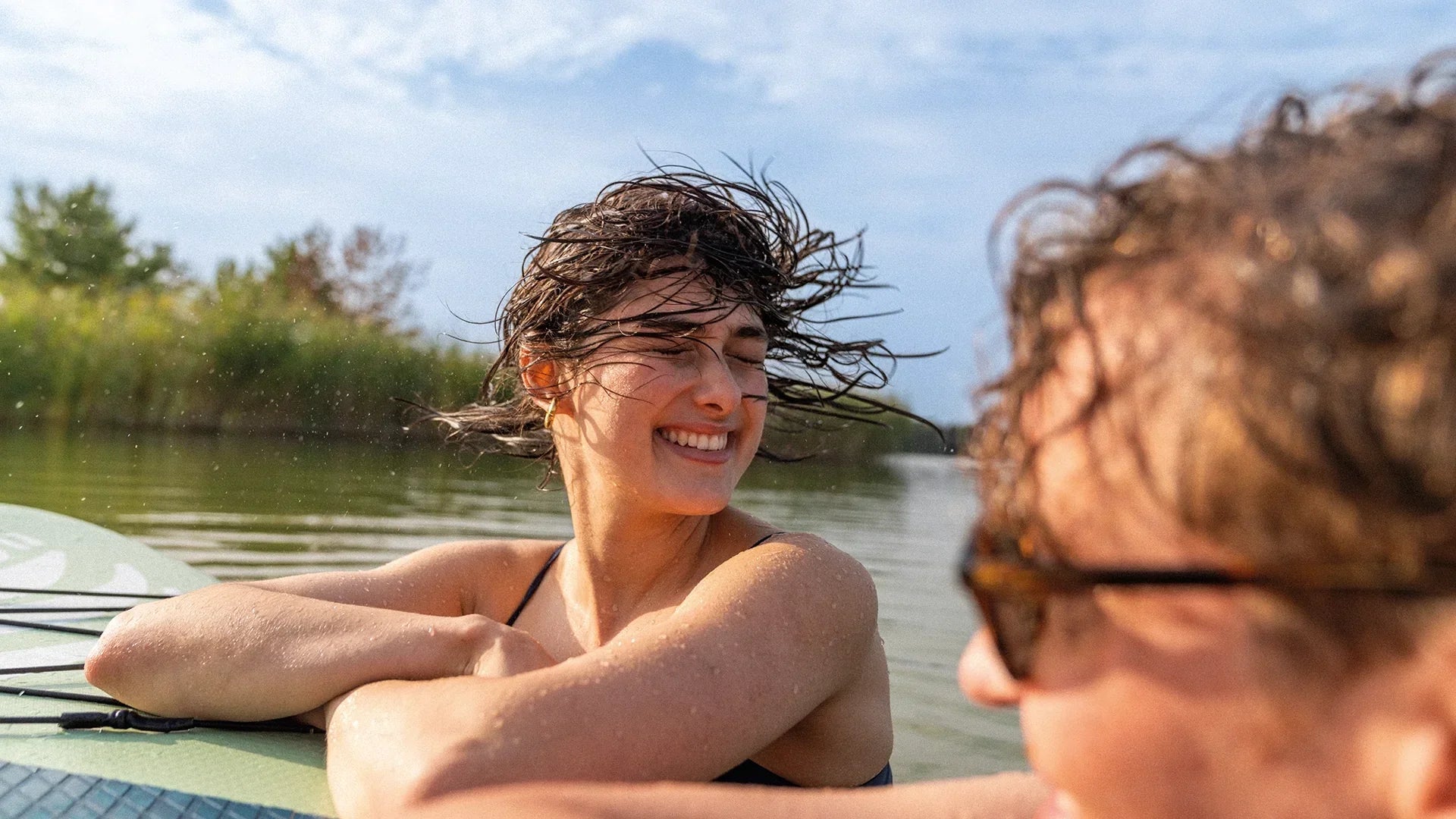 Two people are swimming in a lake with a SUP. The woman shakes her had and waterdrops are flying through the air.