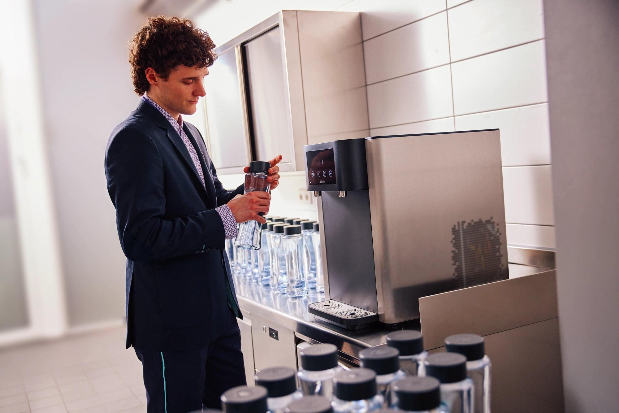 Person using a BRITA Water Dispenser to fill a reusable glass bottle in a kitchen area.