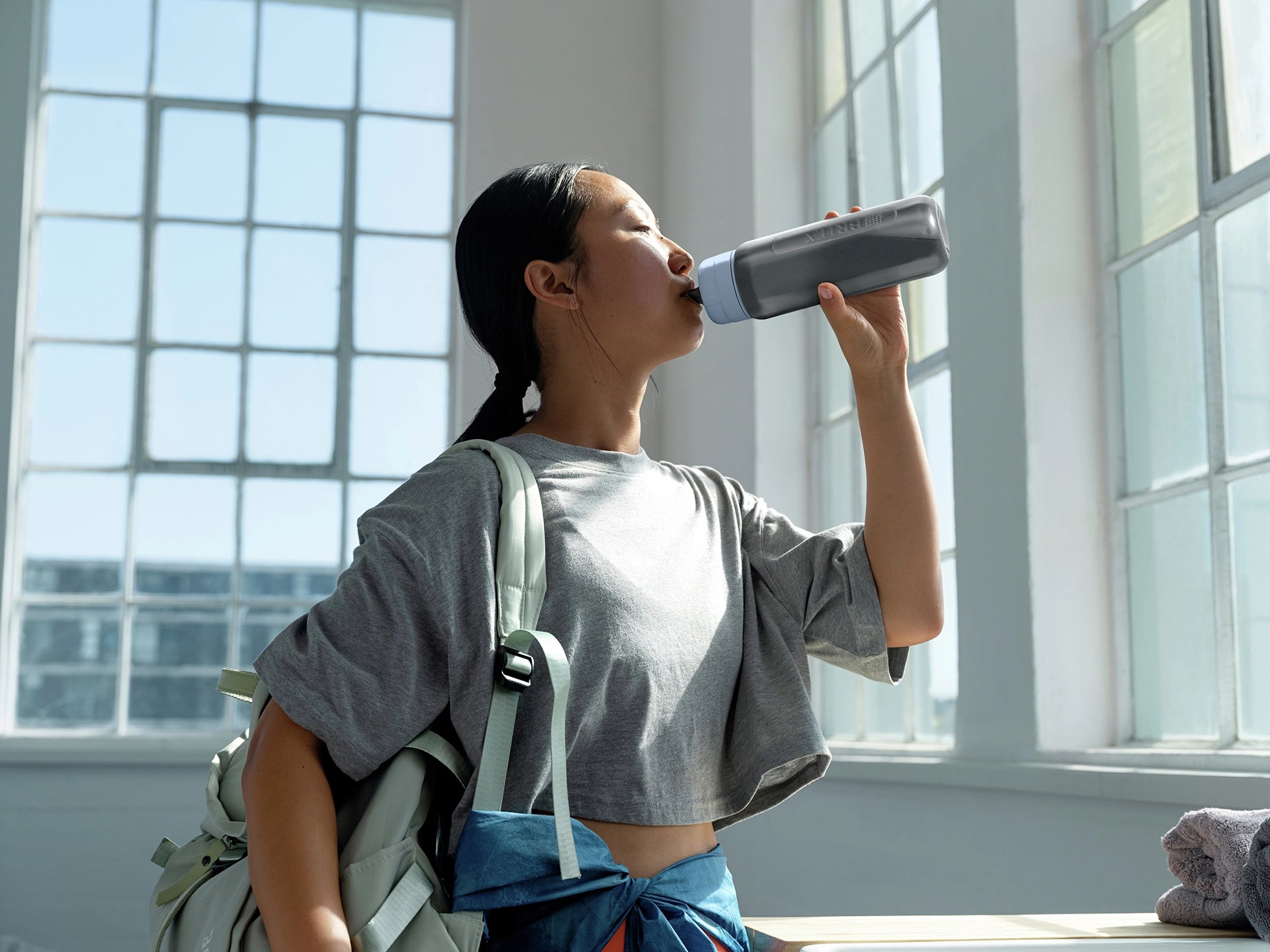 Woman drinking from a BRITA water filter bottle Model Active in a bright room with large windows.