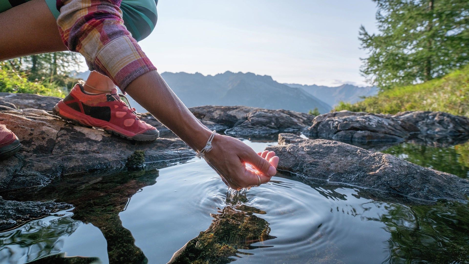 Person touching water in a natural setting with mountains in the background.