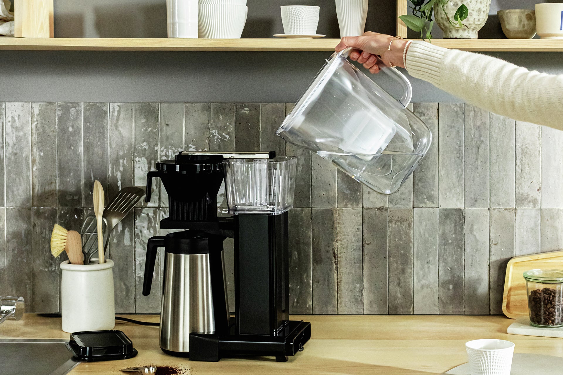 Person pouring filtered water from a white BRITA Style Essential water filter jug into a coffee machine water tank.